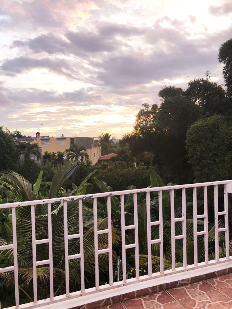 Pastel sunset over palm trees and rooftops seen from the home's white terrace railing